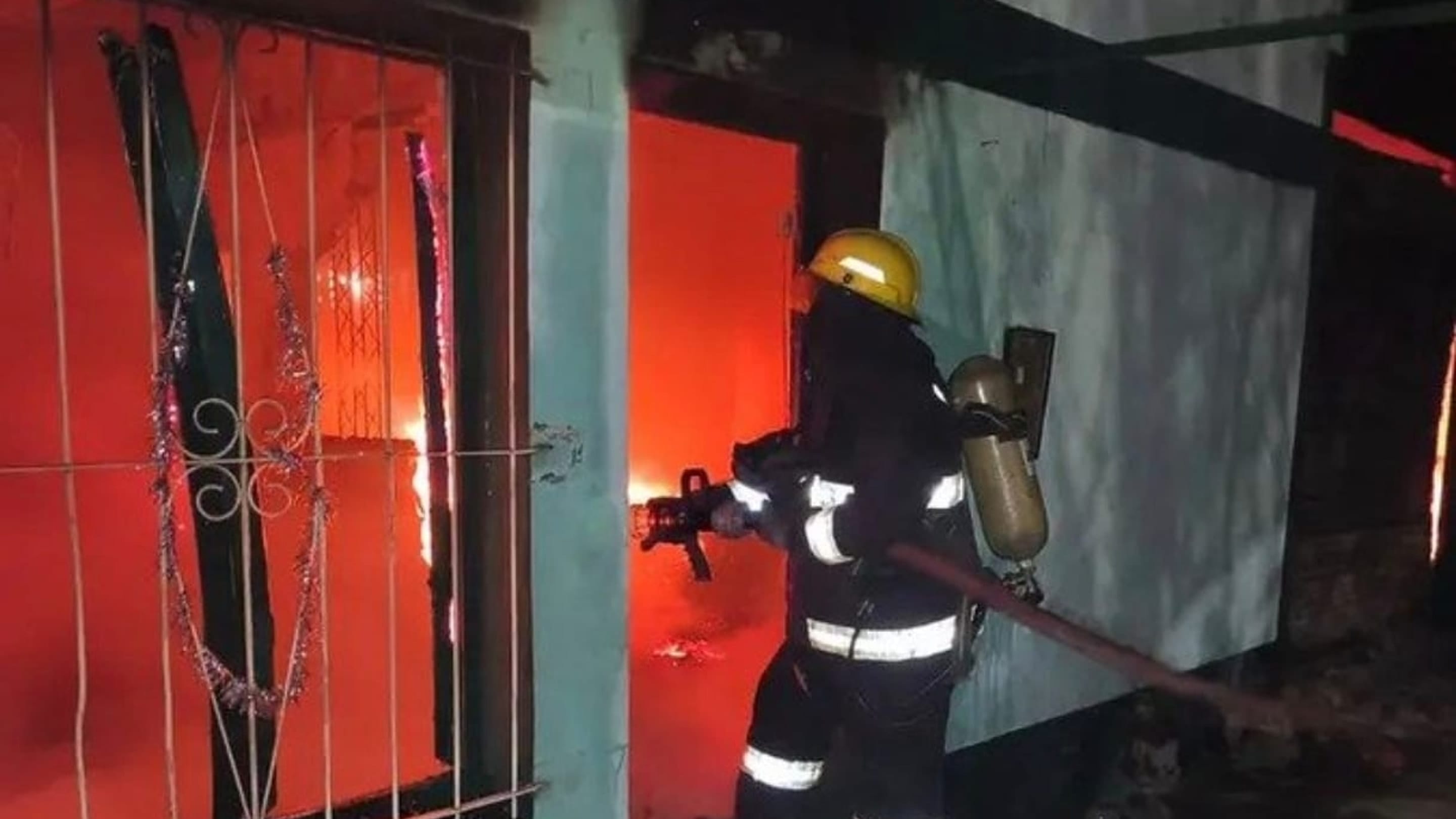 Un bombero con casco y equipo de respiración entra por la puerta de la casa, de noche. La pared es verde agua y por la ventana enrejada se ve el resplandor naranja del fuego adentro.