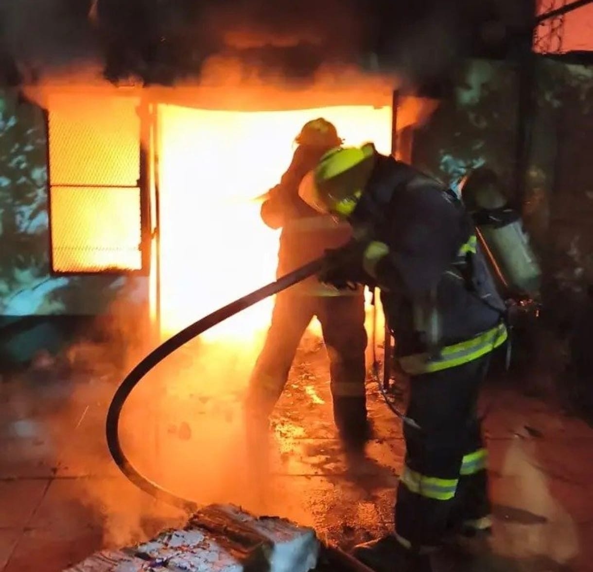 Dos bomberos con manguera frente a una abertura de la que salen llamas altas y humo negro. El agua ya mojó el piso.