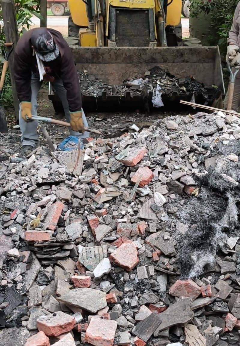 Un hombre con pala junta escombros en un montículo de ladrillos partidos y ceniza. Detrás, la pala de una máquina cargadora amarilla ya está llena de restos quemados.