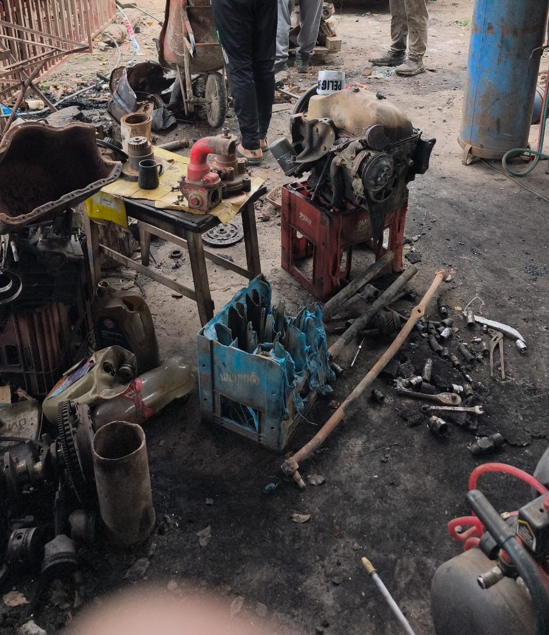 Piezas de motor quemadas apoyadas en el piso y sobre una mesa, ordenadas por tipo: caños, múltiples de escape, un cajón azul con herramientas. Se ven las piernas de varias personas trabajando alrededor.