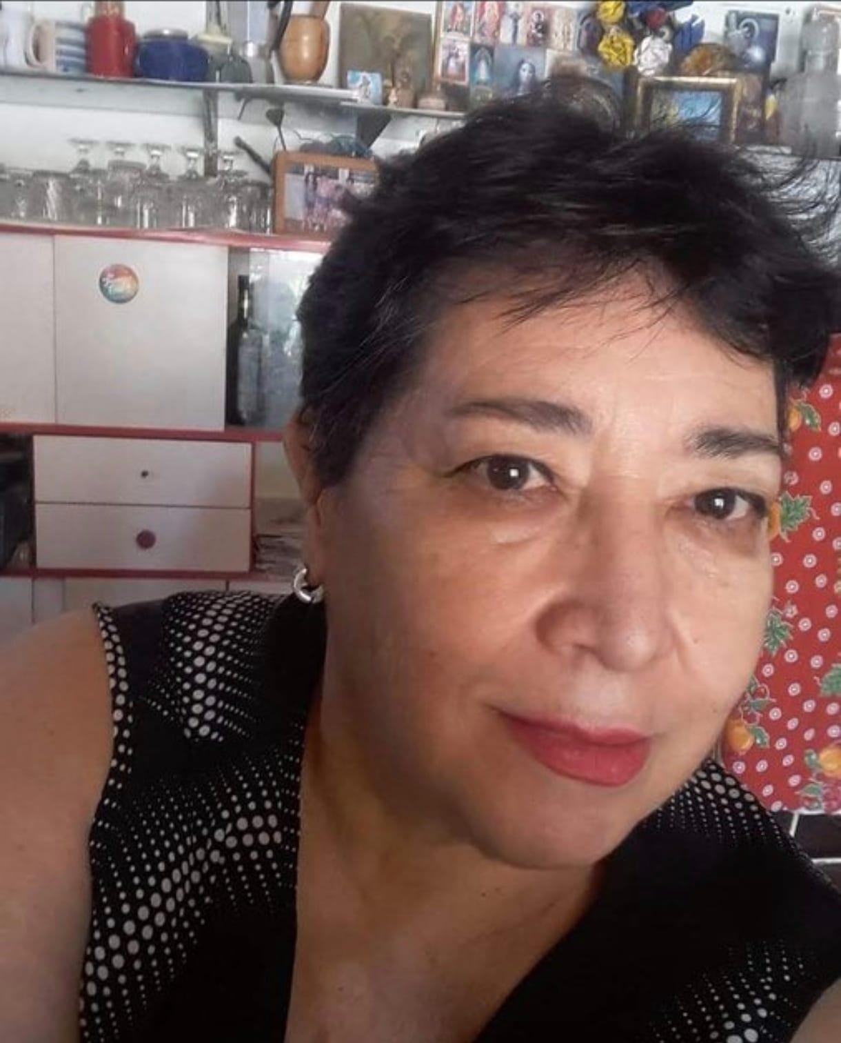 Norma mira a la cámara, sonriendo apenas. Está en su cocina, con una blusa negra de lunares blancos. Detrás hay una alacena con copas y un estante con fotos familiares enmarcadas.